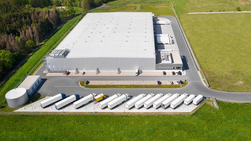 An aerial view of a commercial building with a flat roof and large trucks parked in the parking lot surrounded by a driveway and grass